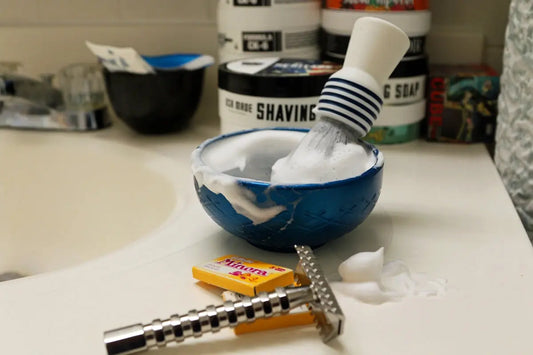 Shaving brush and bowl with lather and razor on sink.